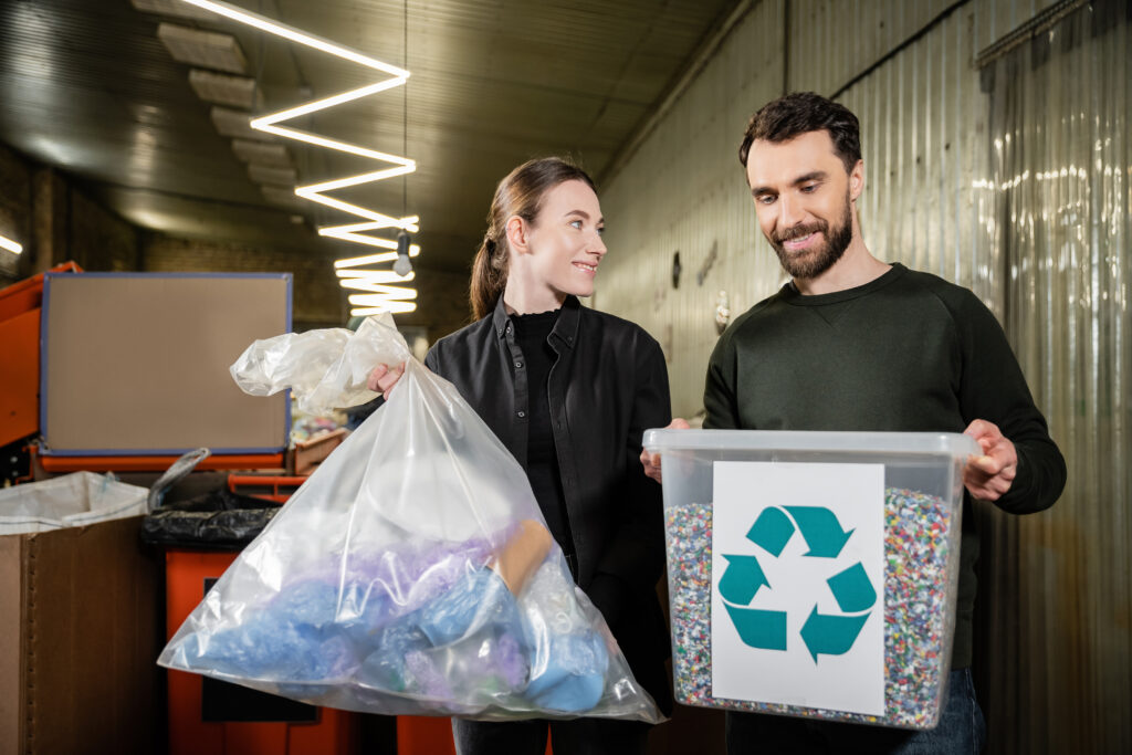 Two people sorting plastic waste at a recycling facility—woman holding a bag of plastic items, man holding a container with recycled plastic pellets. Biobased plastics